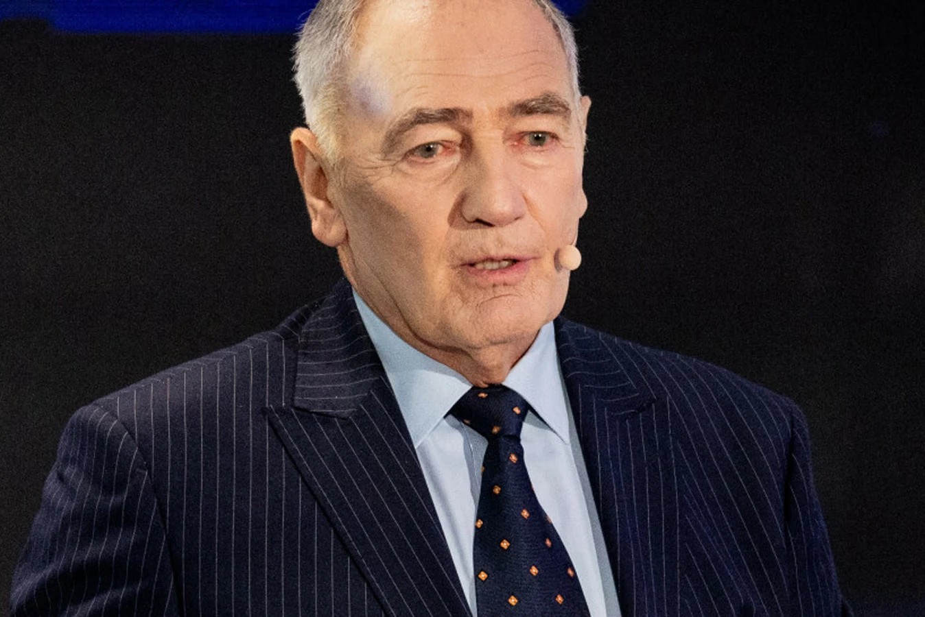 Karol Strasburger Starszy mężczyzna w garniturze i krawacie na tle czarnej sceny, z mikrofonem przypiętym do ucha.