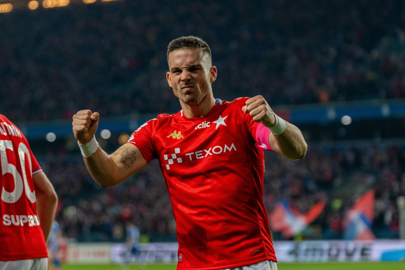 Angel Rodado prowadzi Wisłę Kraków ku Ekstraklasie Piłkarz w czerwonej koszulce drużyny świętuje zaciśniętymi pięściami, tło stanowi stadion z widownią oraz flagami kibiców.