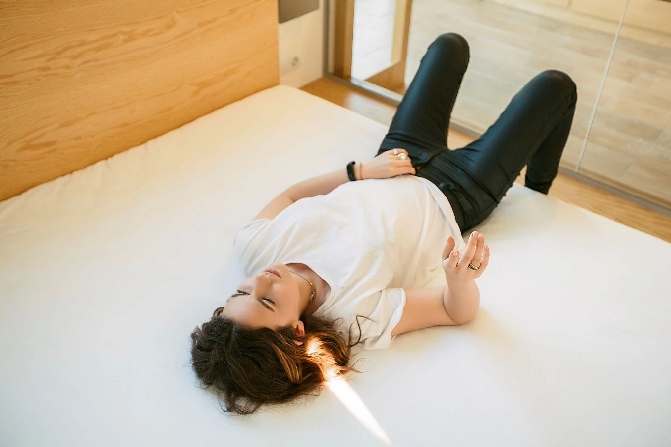 Co oznacza szwajcarski certyfikat? Młoda kobieta w białej koszulce i czarnych spodniach leży na plecach na jasnym, pustym łóżku, rozluźniona, z lekko rozrzuconymi rękami i nogami w minimalistycznie urządzonym, jasnym pokoju.