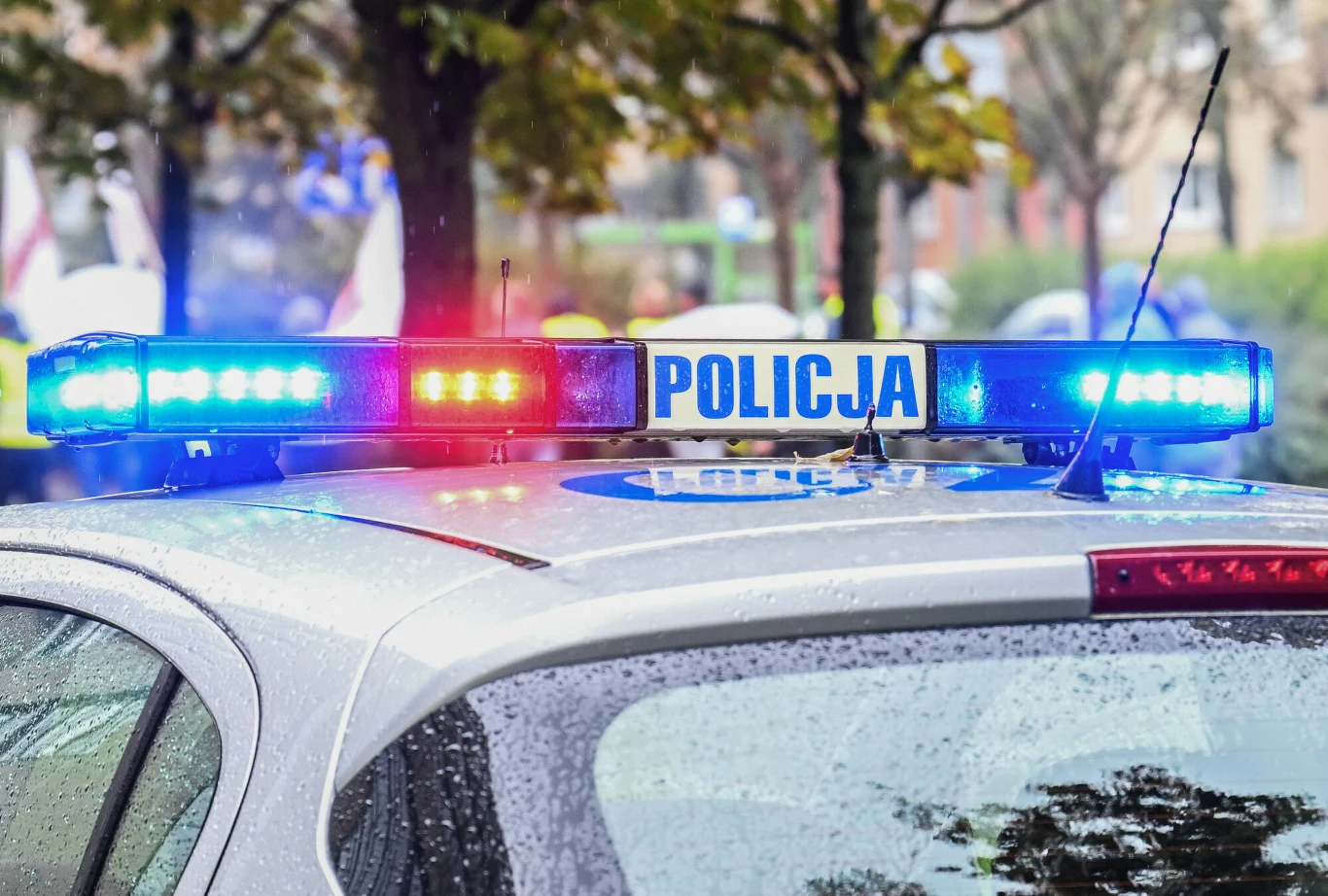 Atak nożownika w Myślenicach. Policjant śmiertelnie postrzelił napastnika. Zdj. ilustracyjne Radiowóz policyjny z włączonymi sygnałami świetlnymi zaparkowany na ulicy, w tle drzewa i nieostre postacie.
