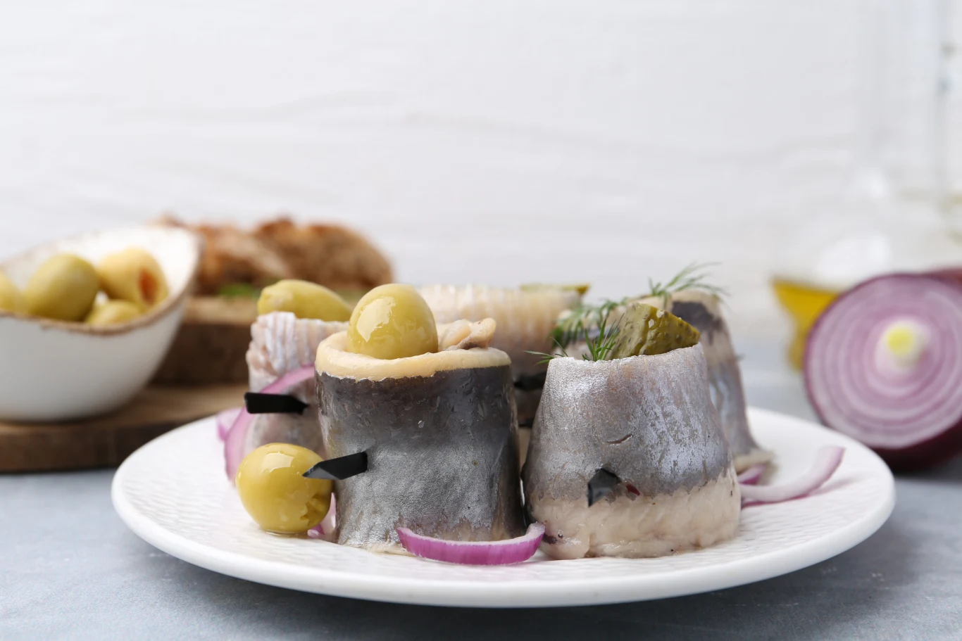 Filety śledziowe zwinięte w rolmopsy, nadziane oliwkami, podane na talerzu z dodatkiem czerwonej cebuli. W tle plaster czerwonej cebuli, oliwki, kromka chleba oraz szklana karafka z oliwą.