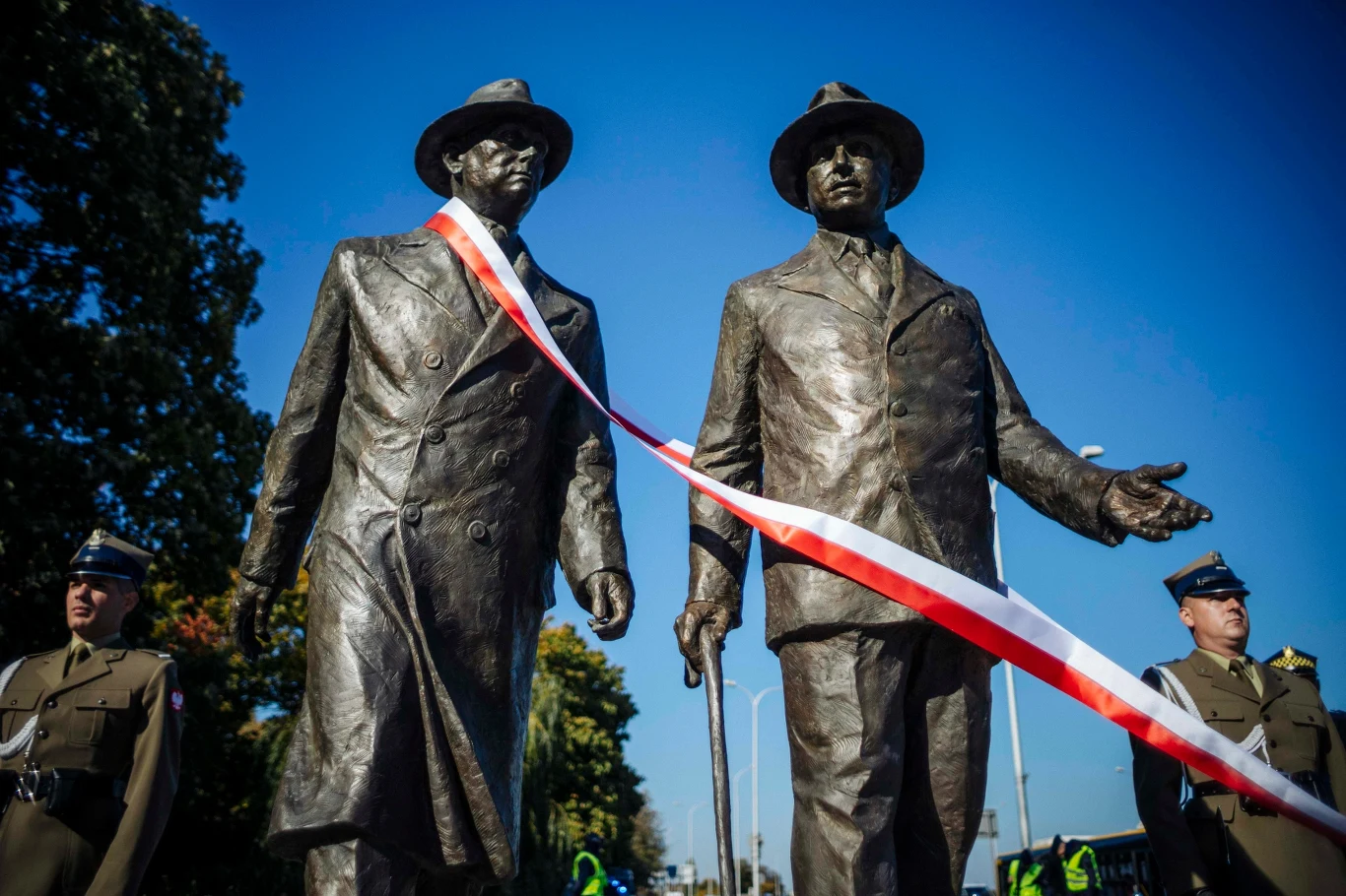 Fundacja Kulskich na rzecz relacji polsko-amerykańskich Dwie postaci z brązu w kapeluszach i płaszczach, spięte biało-czerwoną wstęgą, stoją na tle nieba. Po bokach widać żołnierzy w galowych mundurach.