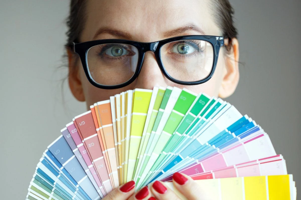 Jaki jest ulubiony kolor osób z wysokim IQ? Kobieta w okularach trzyma rozłożony wachlarz próbek kolorów przed twarzą, skupiając uwagę na palecie barw i jej oczach, które wpatrują się prosto w obiektyw.