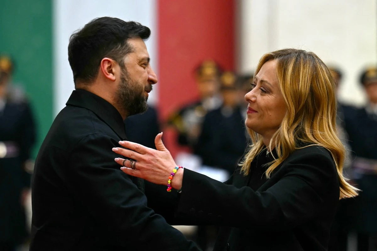 Wołodymyr Zełenski podczas spotkania z Giorgią Meloni Dwoje ludzi wymienia uścisk dłoni, patrząc na siebie z sympatią, na tle rozmytych sylwetek w mundurach oraz dużych pasów w barwach zielonej, białej i czerwonej.