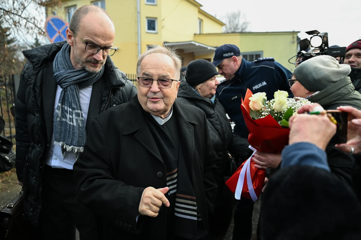 Ojciec Tadeusz Rydzyk stawił się na przesłuchanie Grupa osób zgromadzona na zewnątrz budynku, w centrum starszy mężczyzna w okularach i czarnym płaszczu, obok niego mężczyzna z szalikiem oraz osoby wręczające bukiet biało-czerwonych róż. W tle widoczni policjanci i żółty budynek.