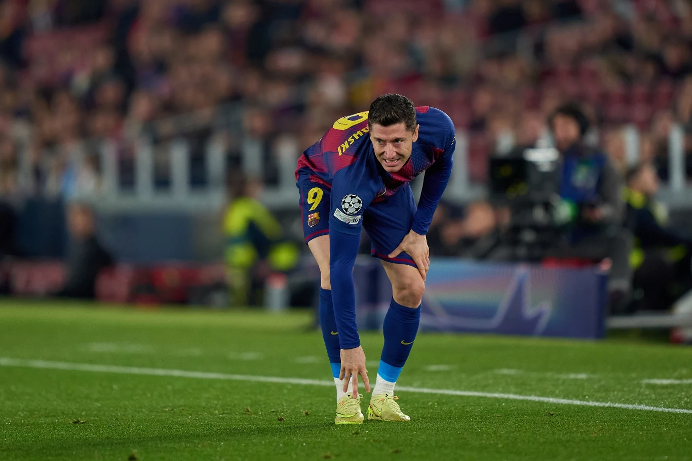 Robert Lewandowski Piłkarz w stroju drużyny FC Barcelona z numerem 9 pochylony na murawie boiska podczas meczu, w tle kibice na trybunach oraz rozmazane sylwetki osób związanych z obsługą spotkania.