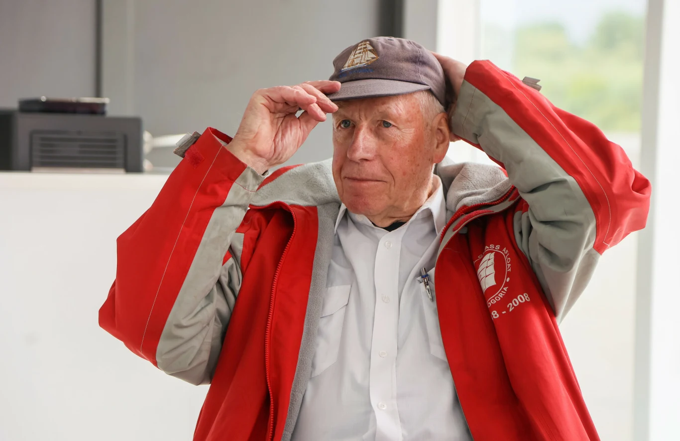 Krzysztof Baranowski w wieku 87 lat zdecydował się na trzeci, samotny rej dookoła świata Starszy mężczyzna w czerwonej kurtce i czapce z daszkiem poprawia nakrycie głowy. Ubrany jest w białą koszulę, patrzy lekko w bok, wydaje się być spokojny i zamyślony.