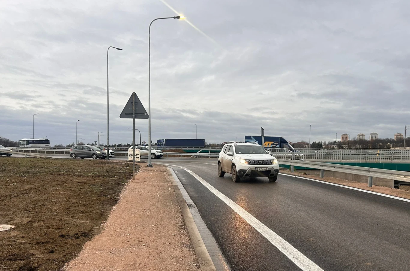 Samochód osobowy jedzie po nowo wybudowanej drodze z asfaltową nawierzchnią, w tle widoczne są inne pojazdy oraz zabudowania, a po bokach barierki ochronne i znak drogowy.