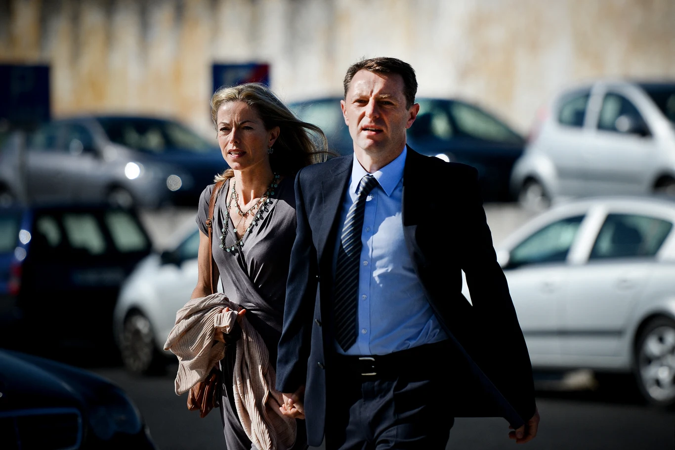 Rodzice Madeleine McCann w 2014 roku. Od lewej: Kate McCann i Gerry McCann Elegancka para dorosłych idzie szybko ulicą, w tle zaparkowane samochody i rozmyta ściana. Mężczyzna ubrany w ciemny garnitur i krawat, kobieta w szarej sukience z torebką w dłoni. Oboje wyglądają na zdeterminowanych lub zatroskanych.