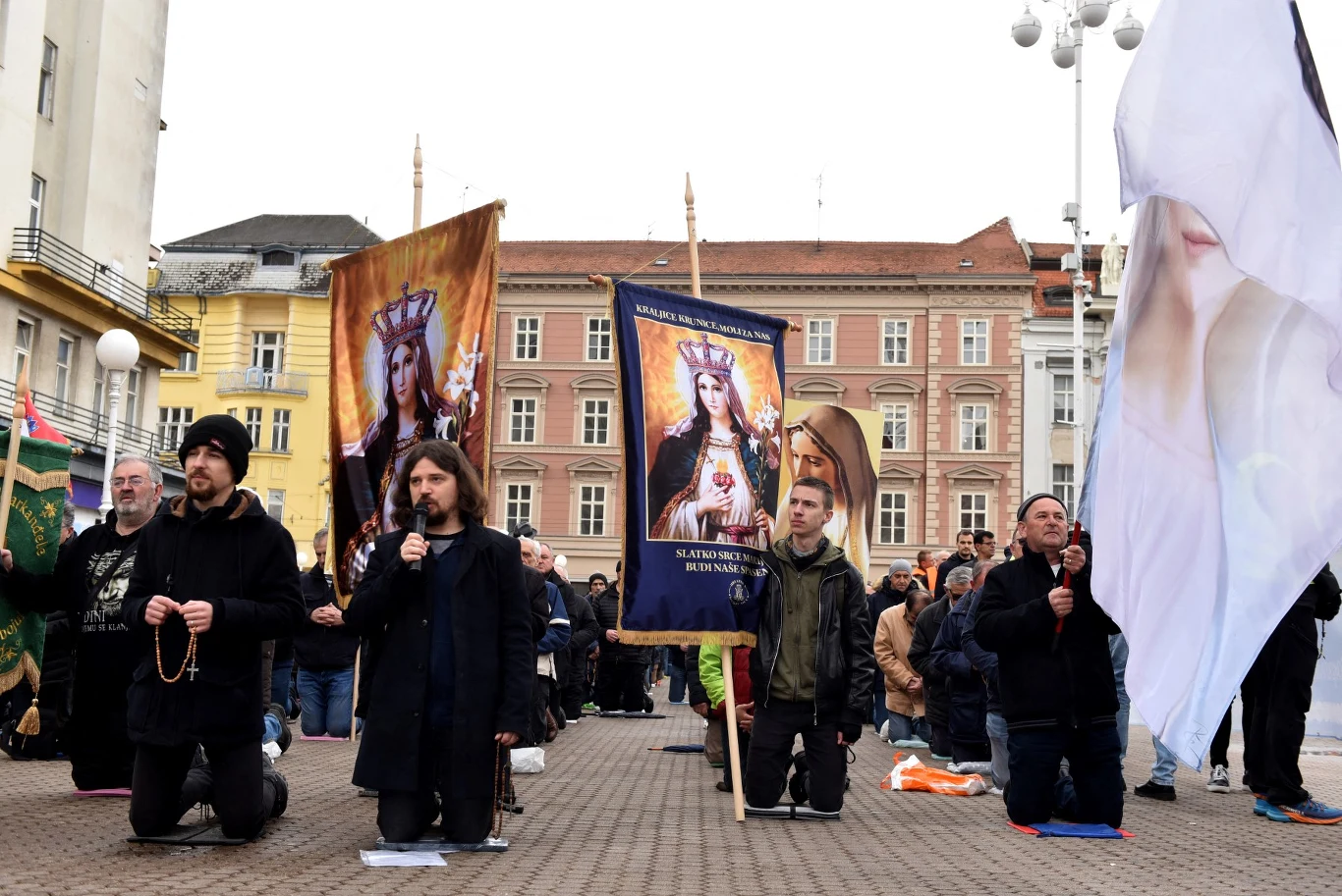 Chorwacja. Ponad 60 tysięcy osób podpisało petycję o usunięcie z przestrzeni publicznej modlitw katolickich mężczyzn promujących patriarchalną władzę Grupa ludzi klęczy na placu miejskim, trzymając banery z wizerunkami świętych i różańce. W tle widoczne są stare kamienice, a wokół zgromadził się tłum uczestników. Atmosfera skupienia oraz religijnego charakteru wydarzenia.