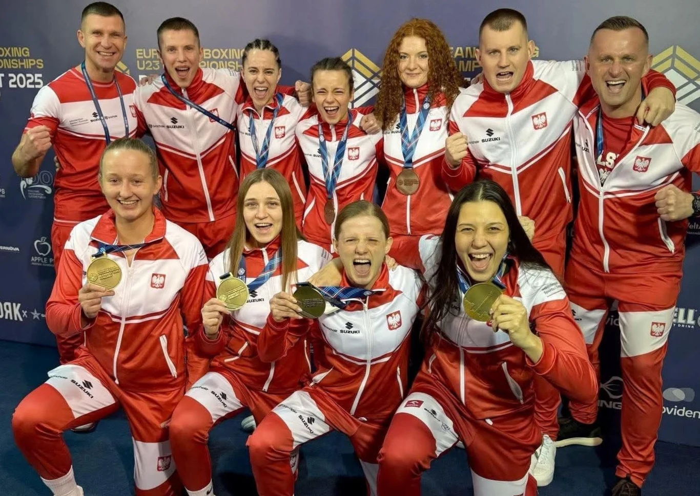 Grupa młodych sportowców w biało-czerwonych strojach z polskim godłem trzymających złote medale i uśmiechających się do obiektywu, na tle ścianki z logotypami mistrzostw bokserskich.