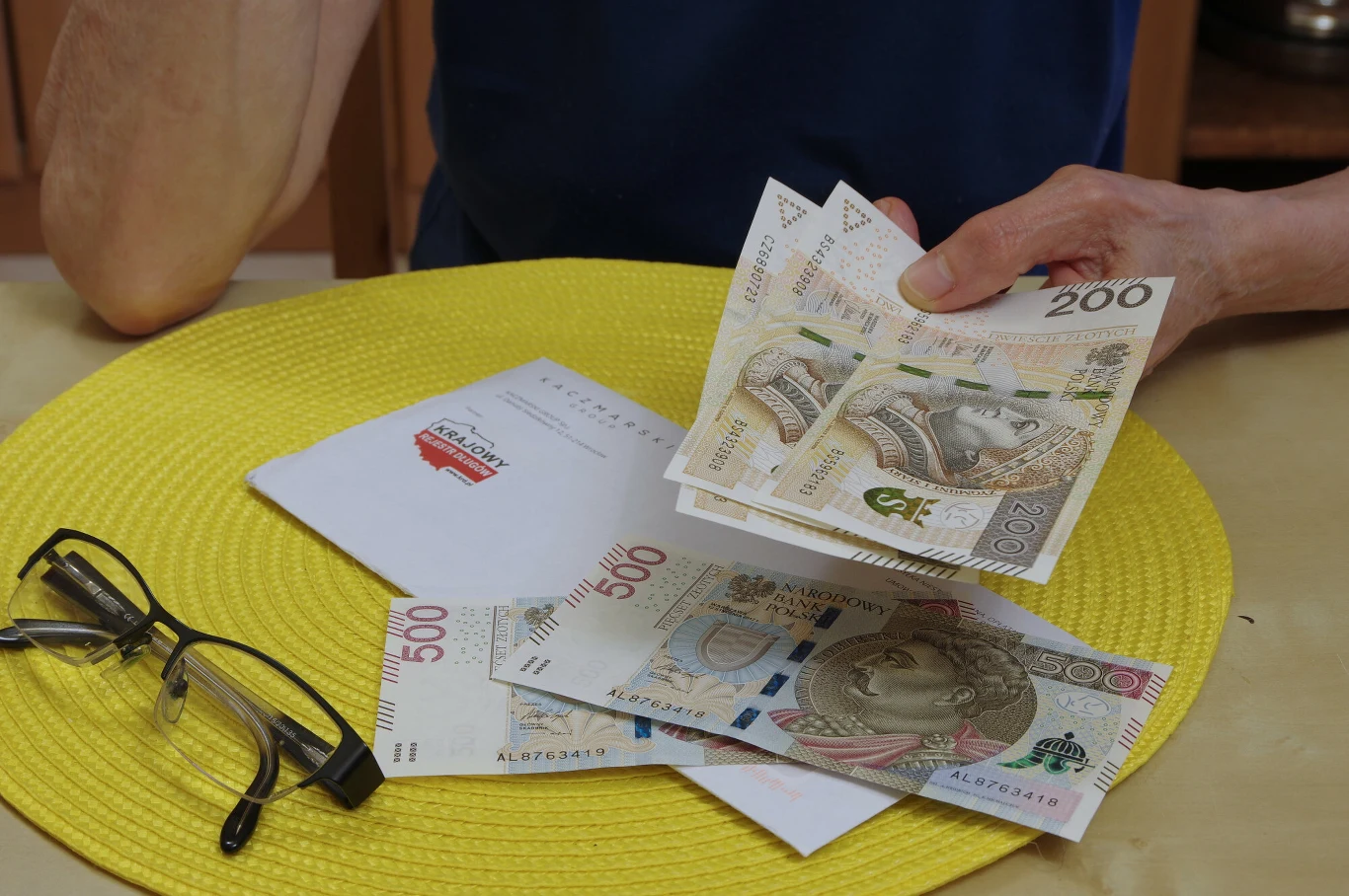 KRD opublikował raport na temat zadłużenia Polaków (zdj. ilustracyjne) Polskie banknoty o nominałach 200 i 500 złotych na żółtej podkładce, obok leży koperta i okulary, osoba w niebieskiej koszuli trzyma część banknotów w dłoni.