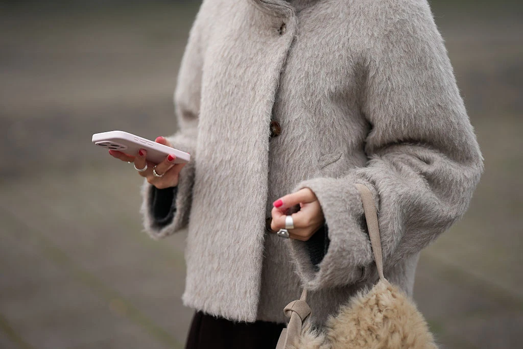 Osoba ubrana w jasny płaszcz z futrzanym elementem trzyma w jednej dłoni jasny smartfon, a w drugiej torebkę z futra. Kolor paznokci jest intensywnie różowy, a na palcach nosi pierścionki.