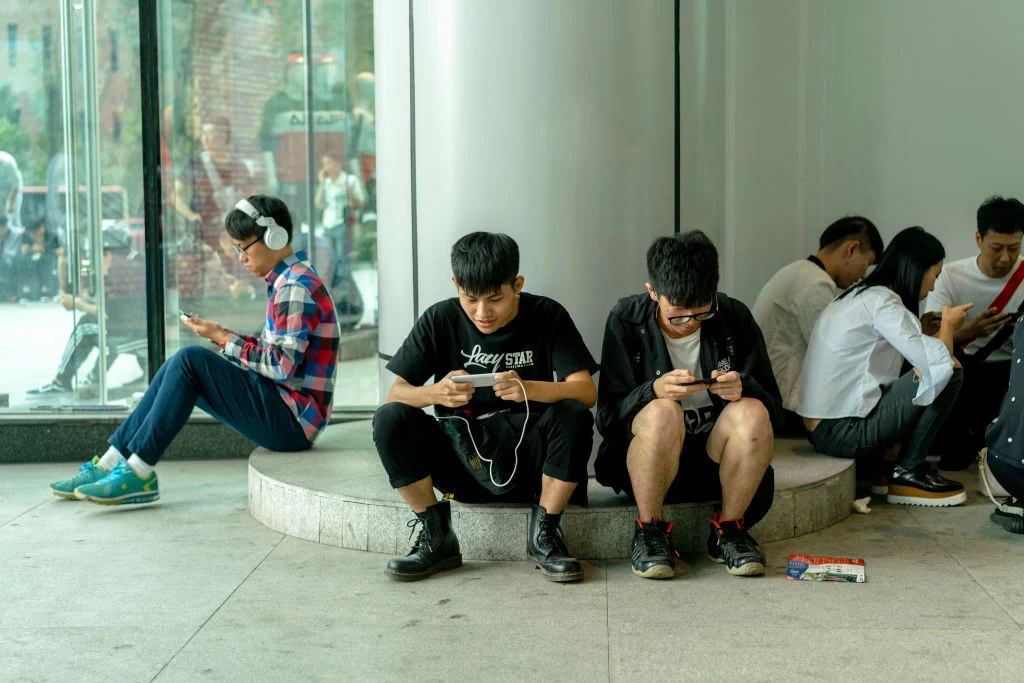 Grupa młodych ludzi siedząca wewnątrz budynku, wpatrzona w ekrany swoich smartfonów, nie zwracając uwagi na siebie nawzajem. Światło dzienne wpada przez duże okno, a otoczenie jest nowoczesne i minimalistyczne.