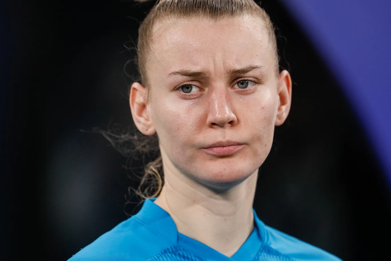 Kobieta o spiętych włosach, ubrana w niebieską koszulkę sportową, z poważnym wyrazem twarzy na tle ciemnego i rozmytego tła.