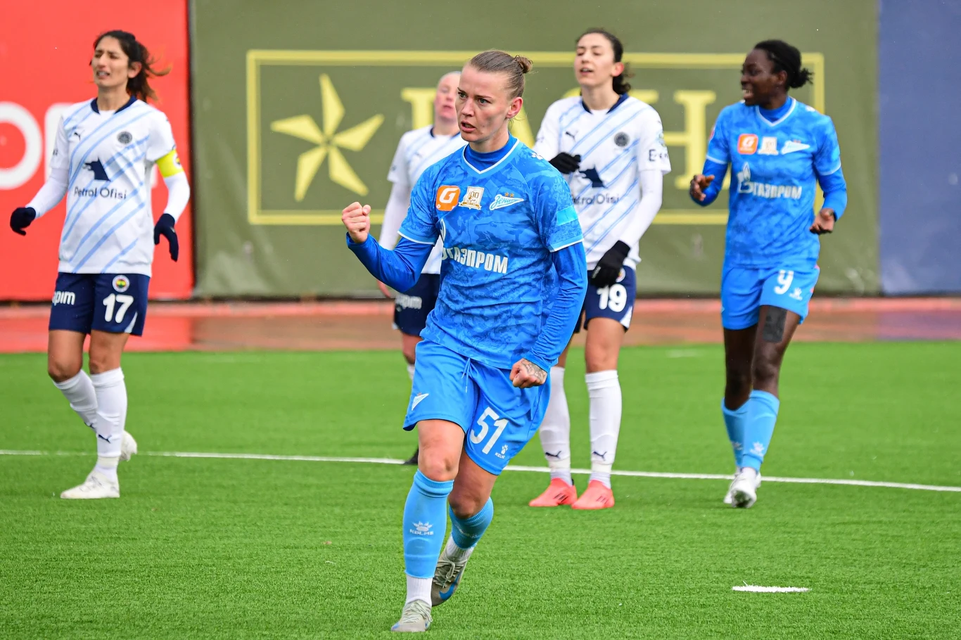 Piłkarka w niebieskim stroju zaciska pięść w geście triumfu podczas meczu piłki nożnej, na tle kilku zawodniczek drużyny przeciwnej ubranych w białe stroje, wszystkie na zielonym boisku.