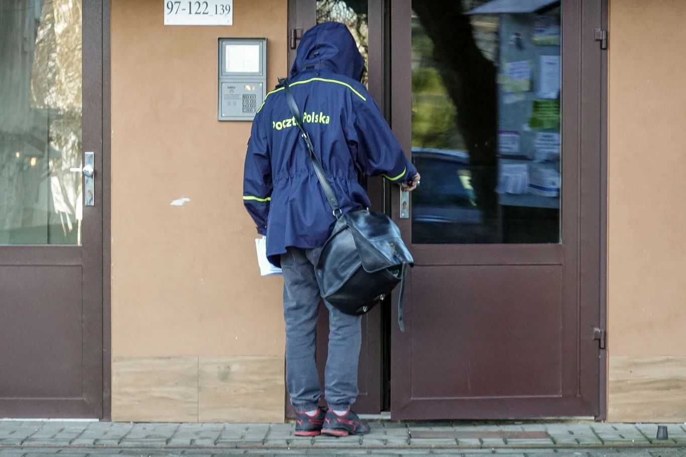 Szukali "fałszywego listonosza", zostali zatrzymani (zdjęcie ilustracyjne) Listonosz ubrany w niebieską kurtkę z napisem 'Poczta Polska' otwiera drzwi do budynku mieszkalnego, trzymając dużą torbę i korespondencję w ręku.