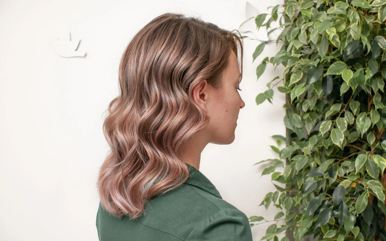 Kobieta o falowanych, jasnoróżowych włosach w zielonej marynarce stoi bokiem, patrząc w stronę dużej zielonej rośliny, tło jasne i minimalistyczne.