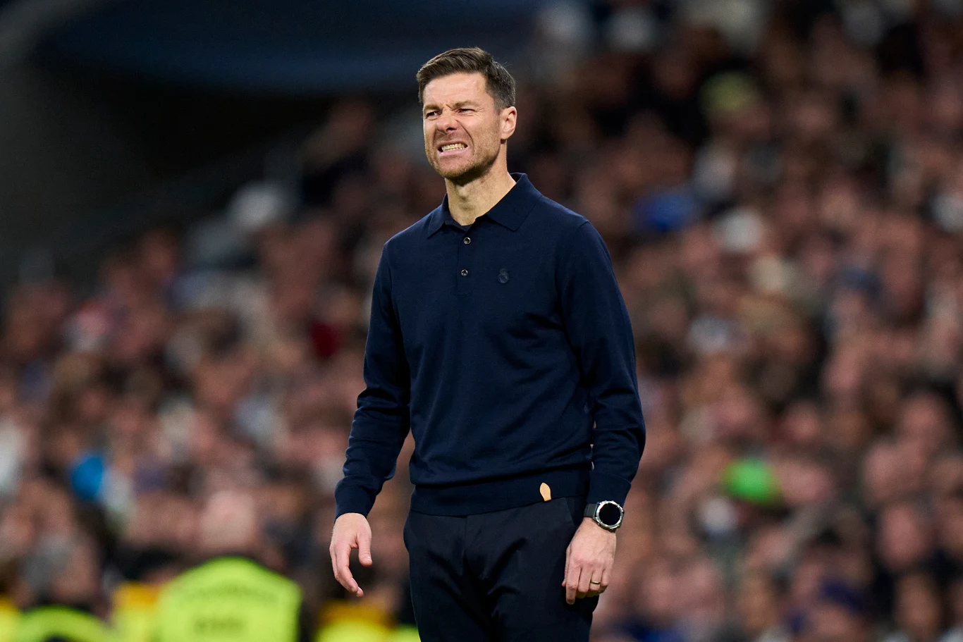 Xabi Alonso Mężczyzna w ciemnym swetrze stojący na tle rozmytego tłumu, z grymasem na twarzy wyrażającym niezadowolenie lub frustrację. Na ręku sportowy zegarek.