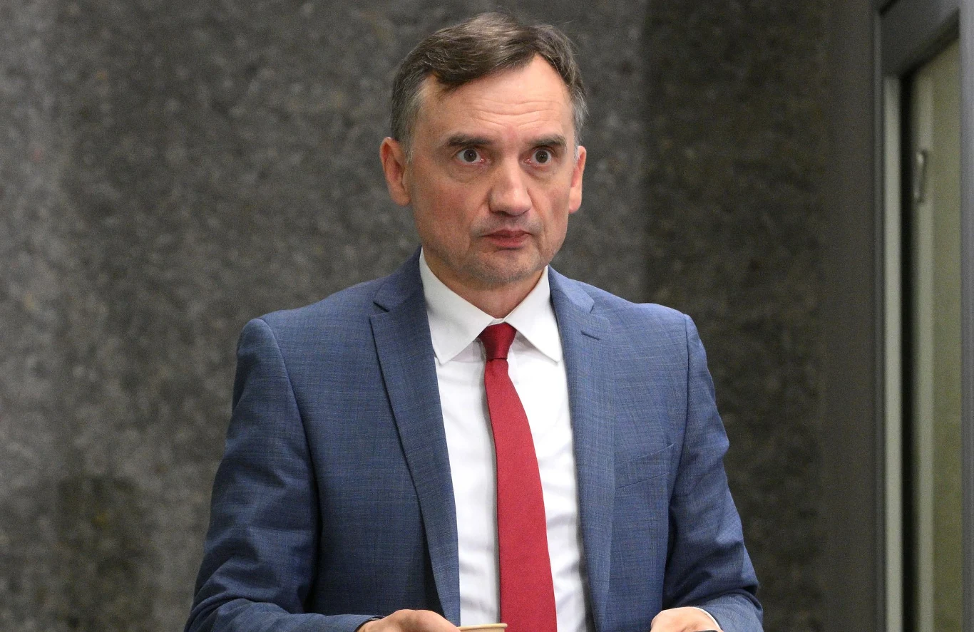 Zbigniew Ziobro Mężczyzna w średnim wieku w eleganckim granatowym garniturze i czerwonym krawacie stoi na tle szarej ściany, trzymając coś w dłoniach.