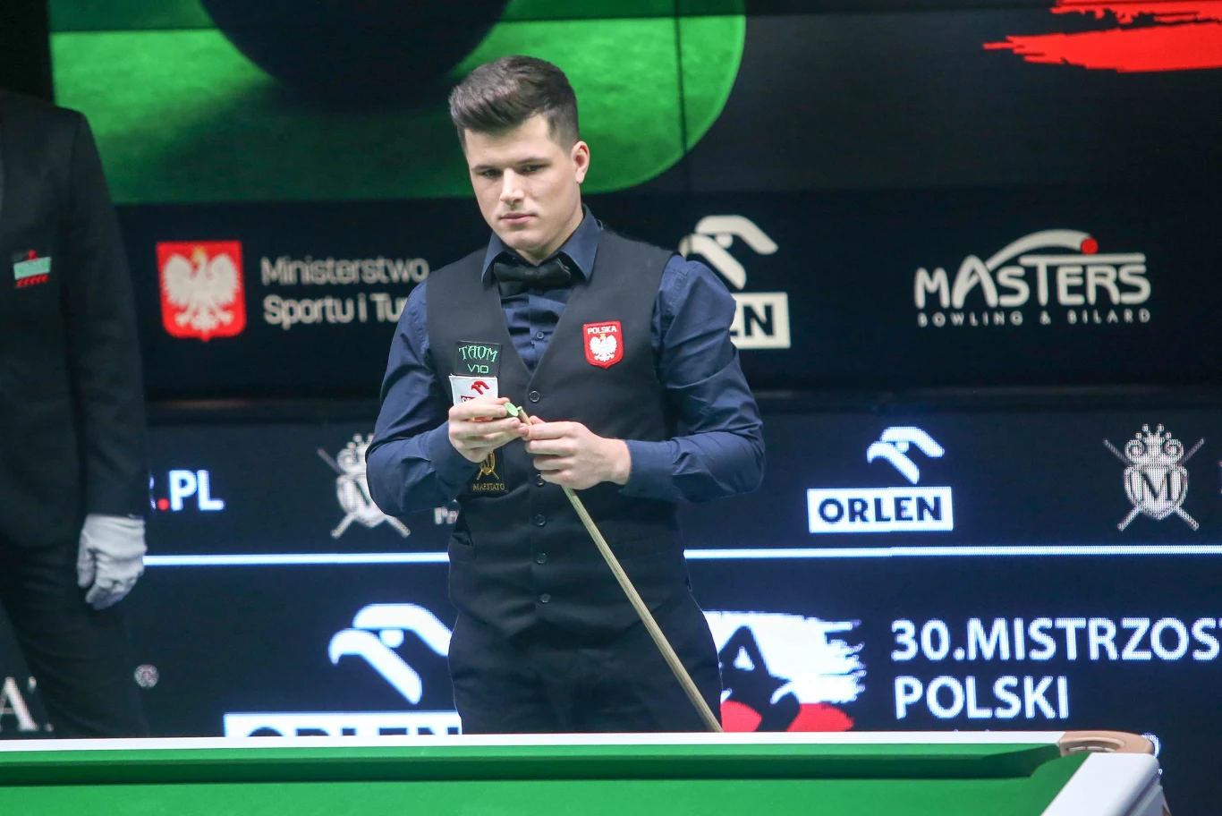 Młody snookerzysta w kamizelce z polskim godłem przygotowuje kij do uderzenia, stojąc przy stole bilardowym na arenie zawodów, w tle widoczne są bannery sponsorów oraz logotypy mistrzostw Polski.