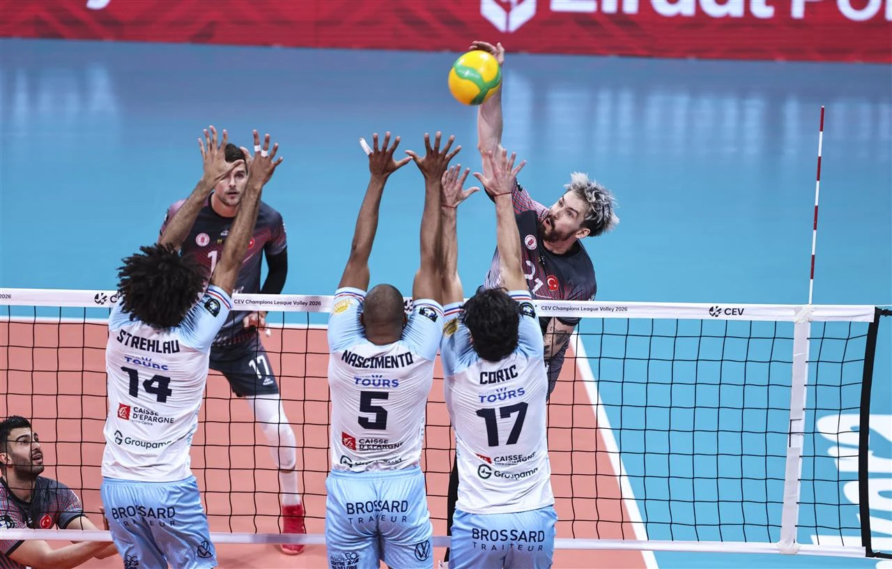 Zawodnik drużyny w ciemnych strojach wykonuje atak nad siatką podczas meczu siatkówki, podczas gdy trzech zawodników drużyny w jasnych koszulkach próbuje zablokować piłkę unosząc ręce nad siatką.