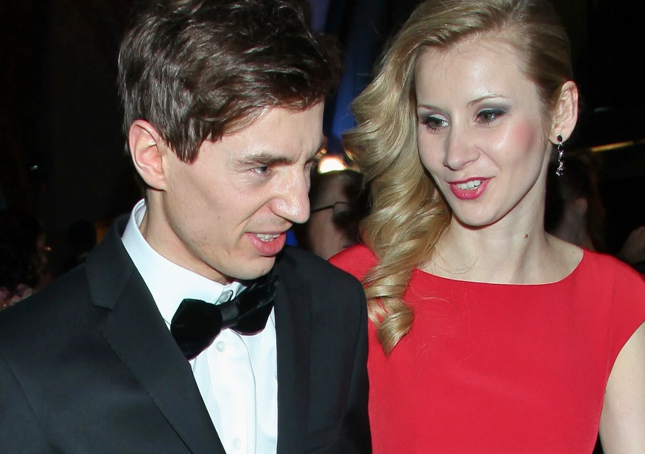 Kamil Stoch, Ewa Bilan-Stoch Para ubrana elegancko, mężczyzna w garniturze z muchą oraz kobieta w czerwonej sukni stoją blisko siebie. Kobieta uśmiecha się i patrzy na mężczyznę, który robi grymas twarzy.