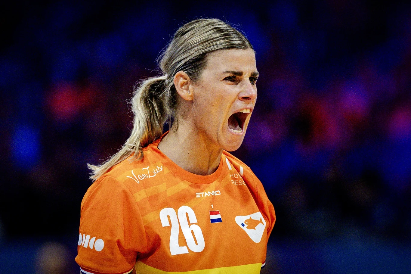 Zawodniczka w pomarańczowej koszulce z numerem 26 głośno wykrzykuje, okazując silne emocje. W tle rozmyte niebieskie światło sugerujące halę sportową. Na koszulce widoczne są logo drużyny oraz flagi Holandii.