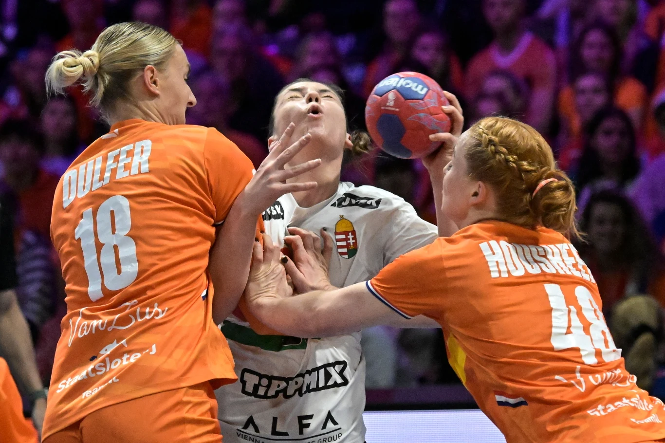 Dynamiczna walka o piłkę ręczną pomiędzy zawodniczką z drużyny w białych barwach a dwiema przeciwniczkami w pomarańczowych strojach, otoczonych przez skupioną publiczność na trybunach.