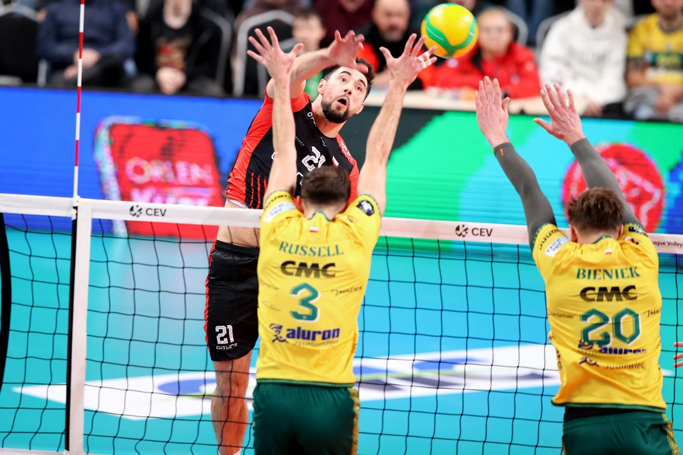 Karol Butryn świetnie rozpoczął mecz z Aluron CMC Wartą Zawiercie Zawodnik w czarno-czerwonym stroju atakuje piłkę siatkową nad siatką, dwóch siatkarzy w żółto-zielonych koszulkach próbuje zablokować piłkę po przeciwnej stronie boiska, tłum kibiców w tle