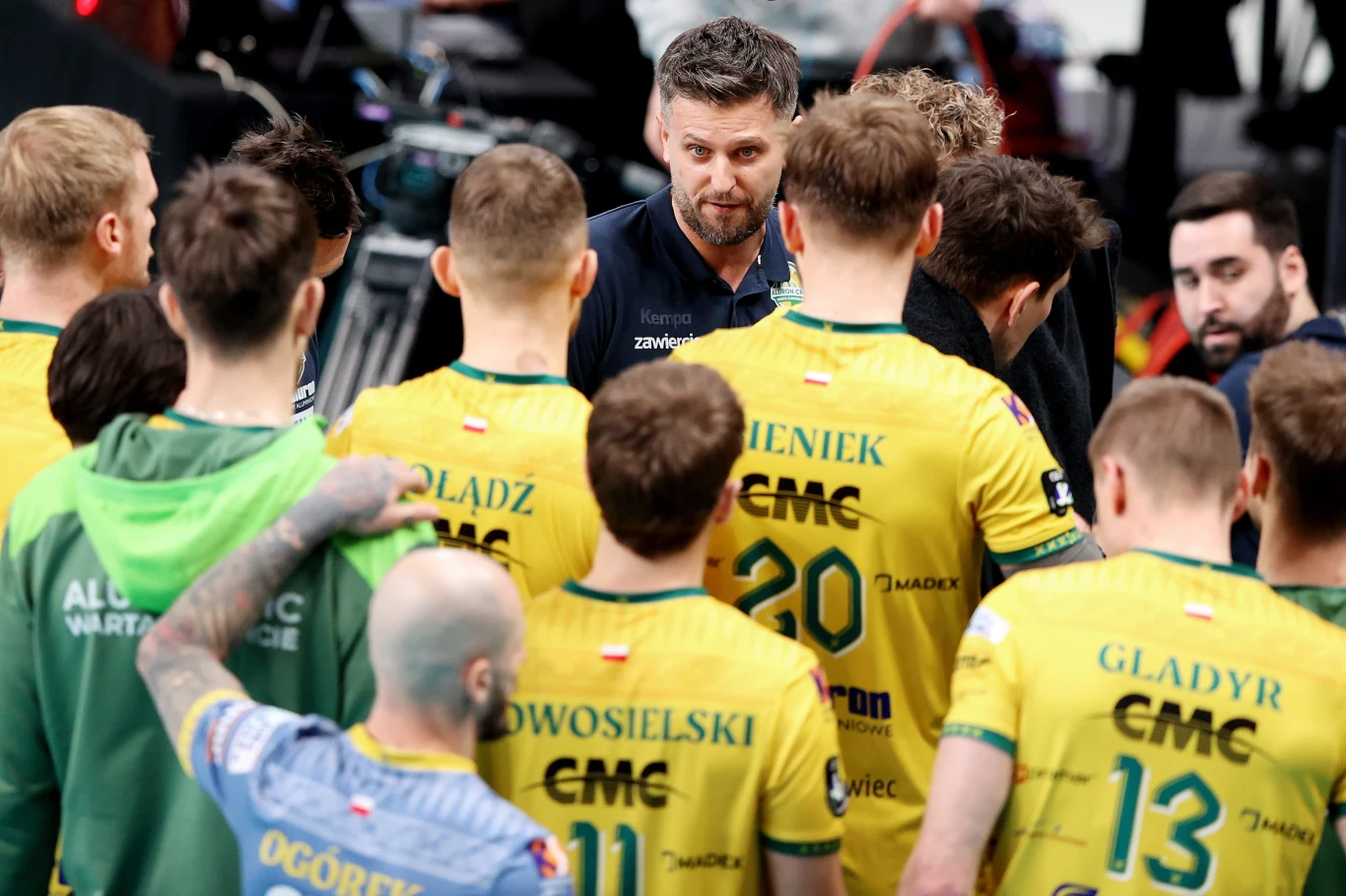 Michał Winiarski i siatkarze Aluron CMC Warty Zawiercie Trener drużyny siatkarskiej omawia strategię z zawodnikami ubranymi w żółte stroje, wszyscy skupieni w kręgu na boisku podczas przerwy w meczu.
