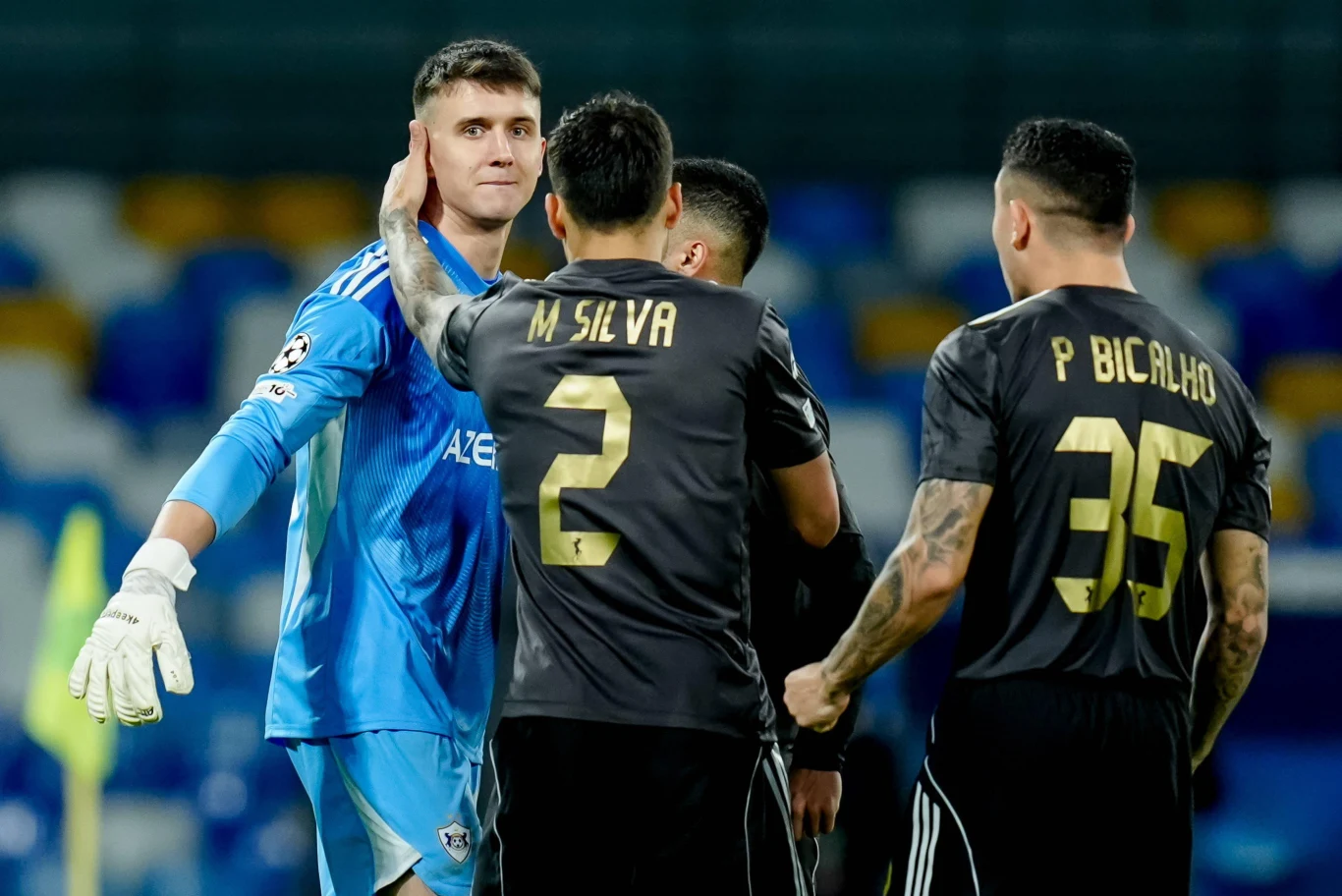 Mateusz Kochalski Grupa piłkarzy podczas meczu, bramkarz w niebieskim stroju podaje rękę zawodnikom w czarnych koszulkach z widocznymi numerami i nazwiskami, na tle trybun stadionu piłkarskiego.