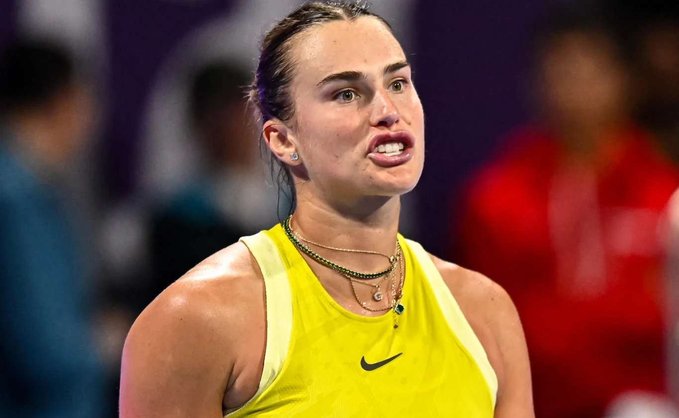 Aryna Sabalenka Kobieta w sportowym żółtym stroju na korcie tenisowym z wyrazem emocji na twarzy. W tle widoczni rozmazani ludzie oraz nieostre światła.
