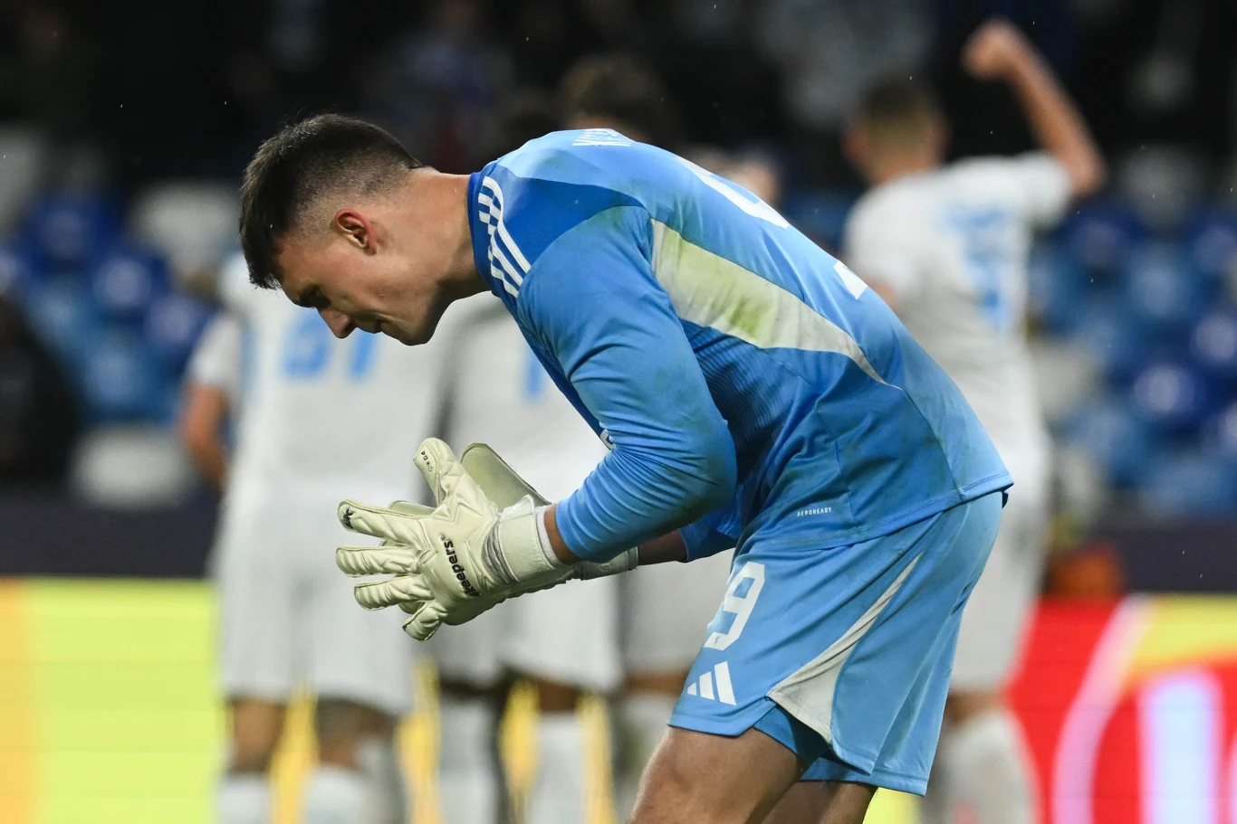 Mateusz Kochalski Bramkarz w niebieskim stroju piłkarskim klęczy na murawie, wyrażając emocje po nieudanym zagraniu, w tle nieostro widoczni pozostali piłkarze oraz trybuny stadionu.