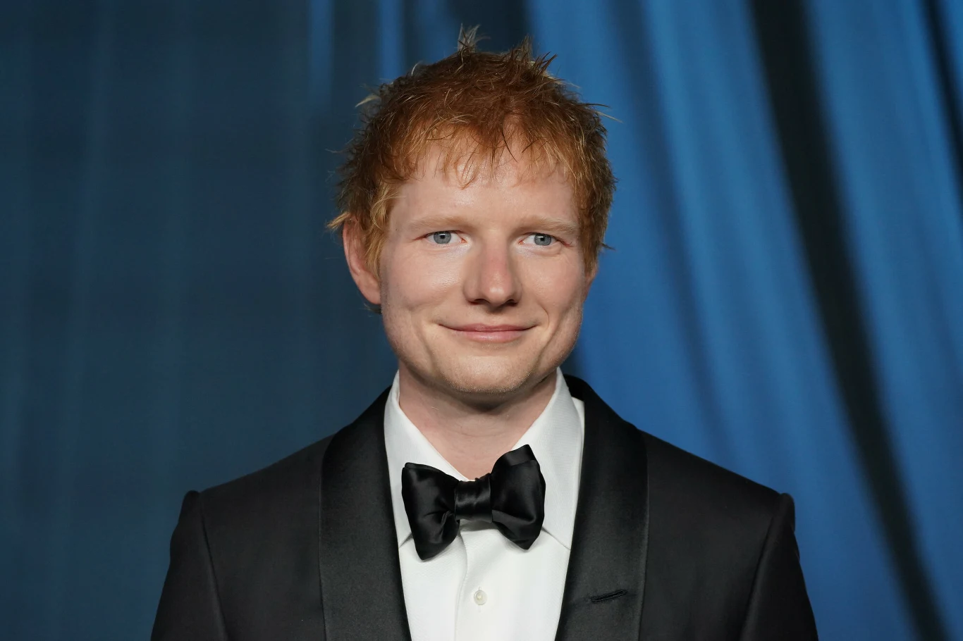 Ed Sheeran Mężczyzna z rudymi włosami i jasną cerą ubrany w czarny smoking z muchą, stoi na tle granatowej, marszczonej tkaniny.
