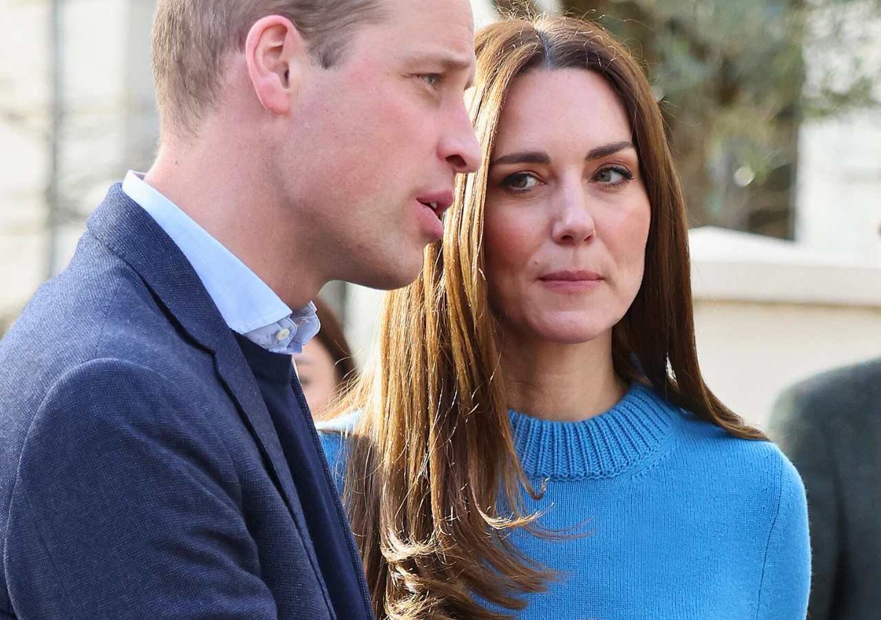 Książę William, księżna Kate Para elegancko ubranych osób w rozmowie na zewnątrz, kobieta w niebieskim swetrze, mężczyzna w marynarce, subtelny kontakt wzrokowy między nimi, za nimi niewyraźne tło z roślinnością.