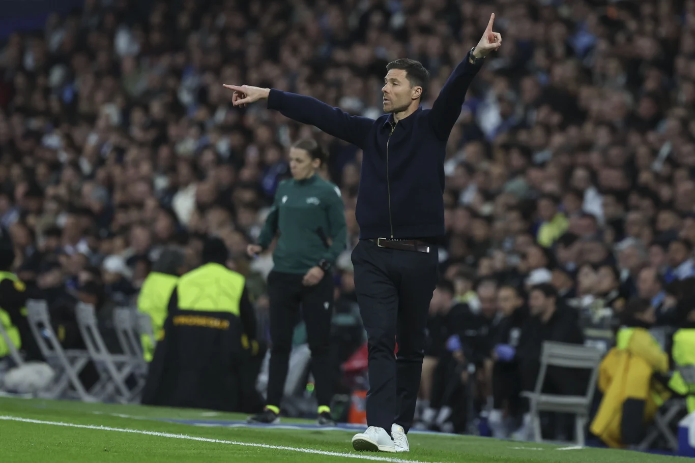 Trener piłkarski energicznie gestykuluje przy linii bocznej podczas meczu, wokół niego widać tłum kibiców oraz osoby zabezpieczające wydarzenie.