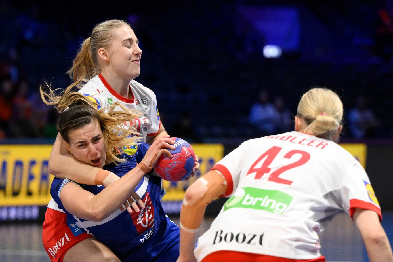 Zawodniczki drużyn piłki ręcznej w dynamicznej akcji podczas meczu, jedna z nich z piłką w rękach, intensywnie broniona przez rywalkę, widoczne emocje i wysiłek sportowy.