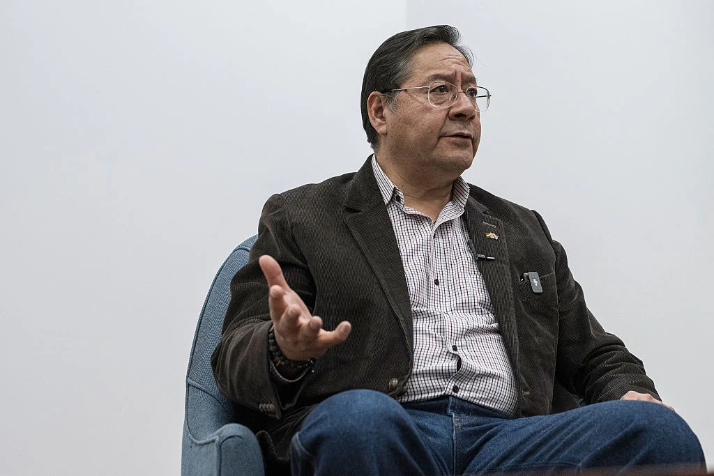 Były prezydent Boliwii Luis Arce Mężczyzna w okularach siedzi na niebieskim fotelu, ubrany w brązową marynarkę i koszulę w kratę, z wyciągniętą ręką w geście wyjaśniania lub rozmowy. Tło jasne, jednolite.