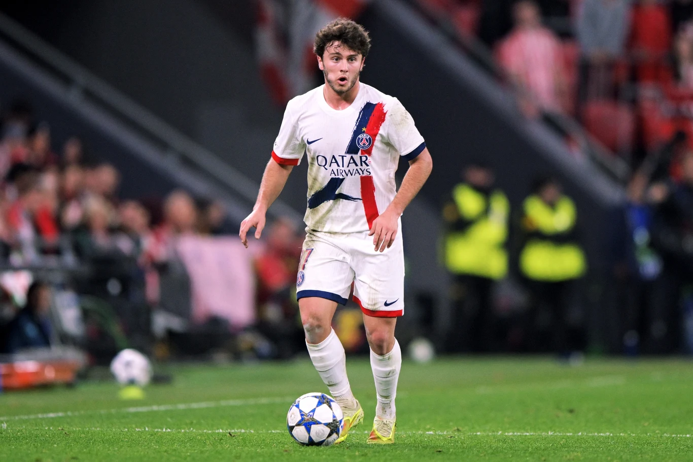 Joao Neves Piłkarz w stroju Paris Saint-Germain biegnie z piłką po murawie stadionu podczas meczu, w tle widoczni kibice oraz ochroniarze.