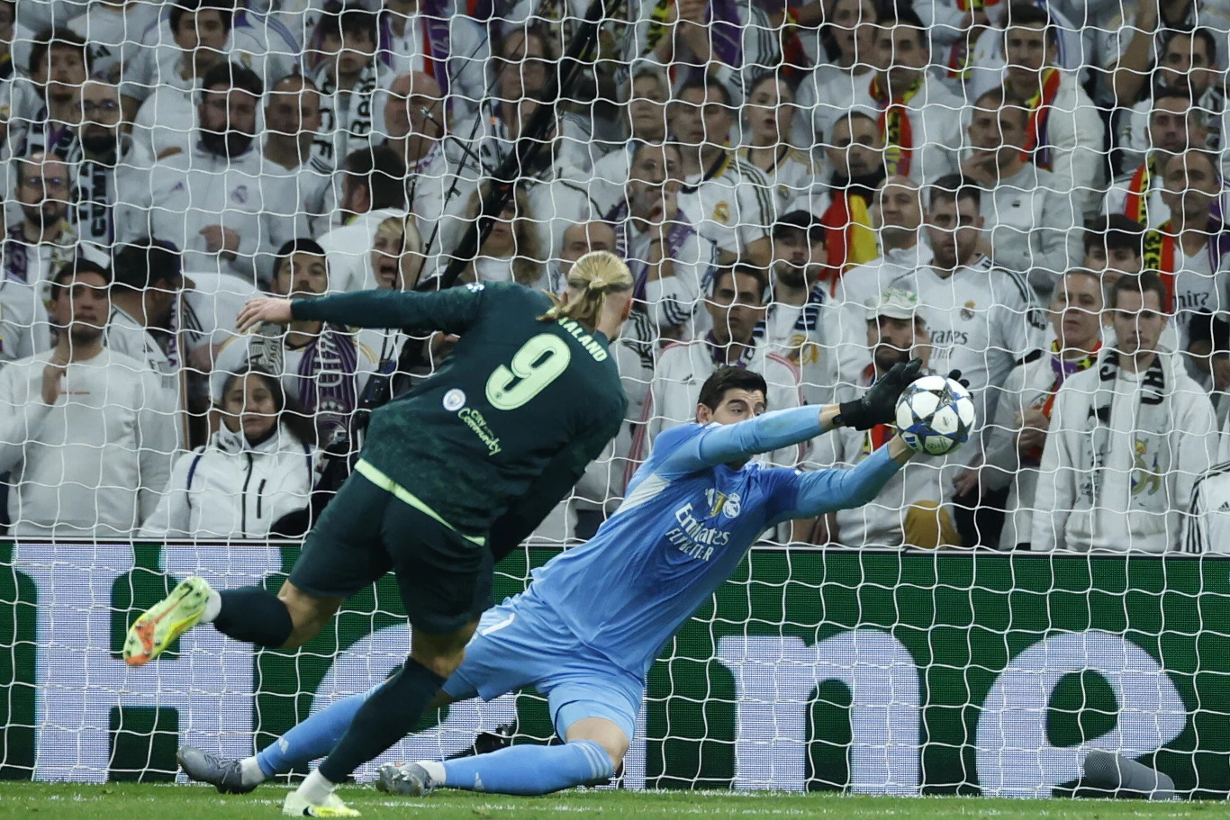 Zawodnik drużyny atakującej próbuje zdobyć bramkę, oddając strzał głową, podczas gdy bramkarz rywali wykonuje efektowną paradę, broniąc piłkę przed wpadnięciem do siatki. W tle trybuny wypełnione kibicami ubranymi w białe stroje.