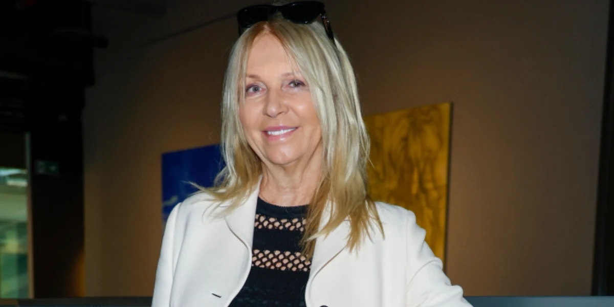 Mariola Bojarska-Ferenc Blondynka w średnim wieku z długimi prostymi włosami ubrana w białą marynarkę oraz czarną bluzkę z siateczkowymi wstawkami uśmiecha się delikatnie, w tle fragmenty obrazów na ścianie.