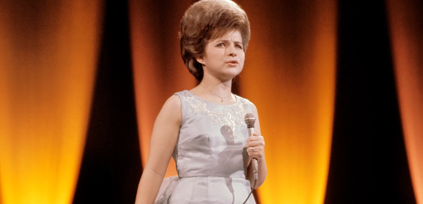 Brenda Lee Młoda kobieta w eleganckiej sukni stoi na scenie z mikrofonem w ręku, tło podświetlone pomarańczowymi i żółtymi światłami tworzącymi efektowną atmosferę występu.