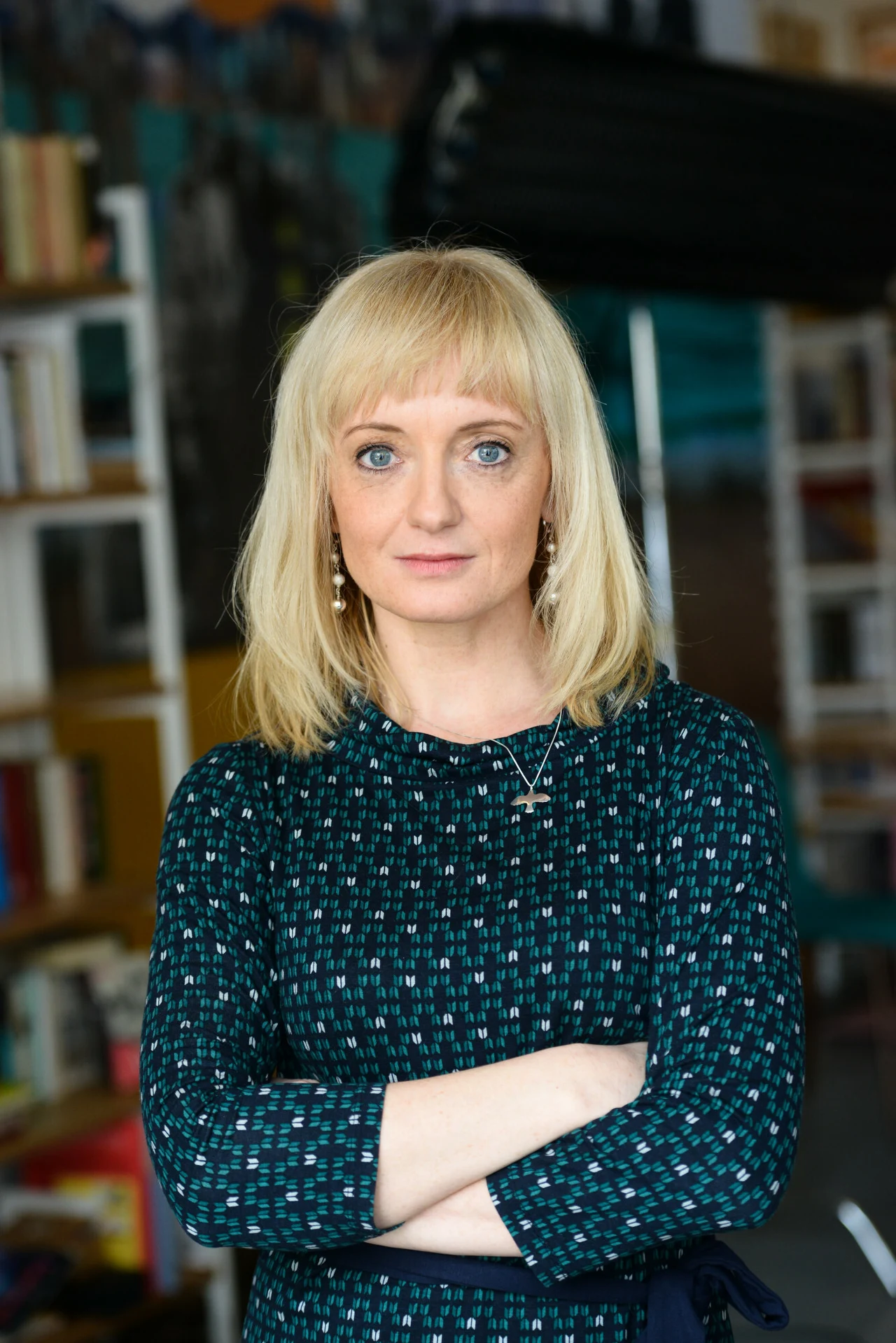 Kobieta o blond włosach i niebieskich oczach stoi z założonymi rękami, ubrana w ciemnozieloną sukienkę w jasne wzory, patrzy pewnie w obiektyw. W tle rozmyte półki z książkami i regały.