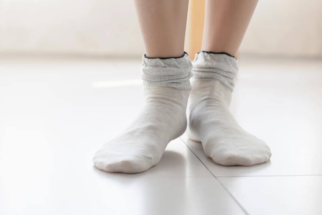 Stopy ubrane w jasne skarpetki stojące na jasnej, gładkiej podłodze w naturalnym świetle.