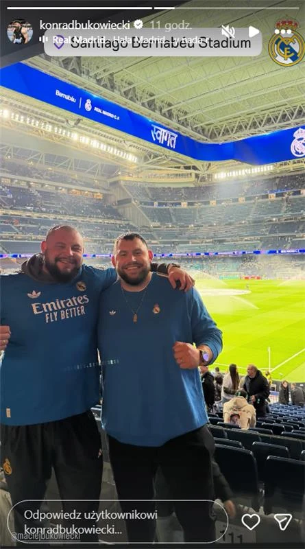 Maciej Bukowiecki i Konrad Bukowiecki na Santiago Bernabeu przed meczem Realu Madryt Dwóch uśmiechniętych mężczyzn w niebieskich bluzach z herbem klubu Real Madryt pozuje na tle trybun stadionu Santiago Bernabéu. W tle widoczna boisko piłkarskie oraz logo klubu na ekranach otaczających trybuny.