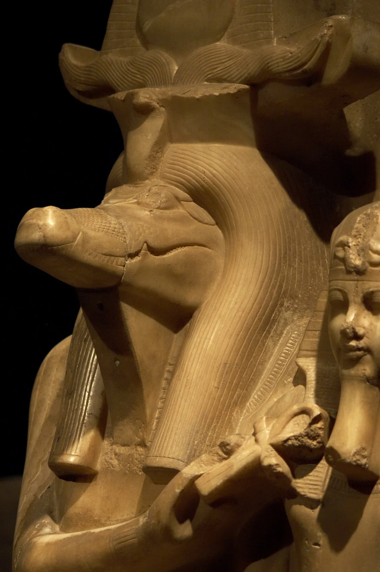 Sobek to egipski bóg przedstawiany z głową krokodyla Starożytna rzeźba z Egiptu przedstawiająca postać z głową krokodyla, prawdopodobnie wizerunek Sobka, egipskiego boga wody, z widocznym fragmentem innej postaci po prawej stronie.