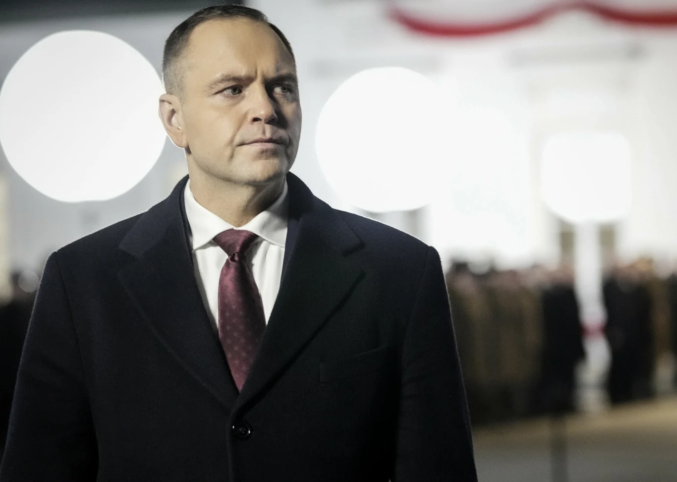 Prezydent Karol Nawrocki Mężczyzna w ciemnym płaszczu i bordowym krawacie stoi na tle rozmytych sylwetek ludzi oraz jasnych świateł, atmosfera jest oficjalna i podniosła.