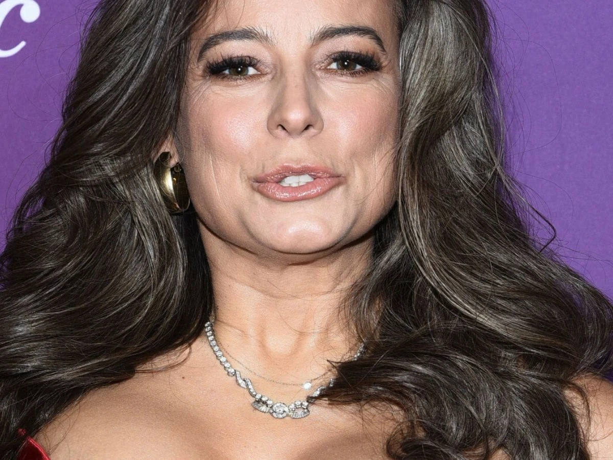 Anna Mucha Kobieta o długich falowanych włosach, w eleganckiej biżuterii, na fioletowym tle. Ubrana w czerwoną suknię, z subtelnym makijażem i wyrazistym spojrzeniem.