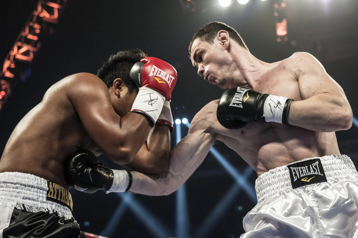 Dwóch bokserów w intensywnej walce, jeden z nich wykonuje mocny prawy cios w głowę przeciwnika. Obaj zawodnicy mają na sobie rękawice i sportowe spodenki. Jasne oświetlenie sceny podkreśla dynamiczny charakter pojedynku.